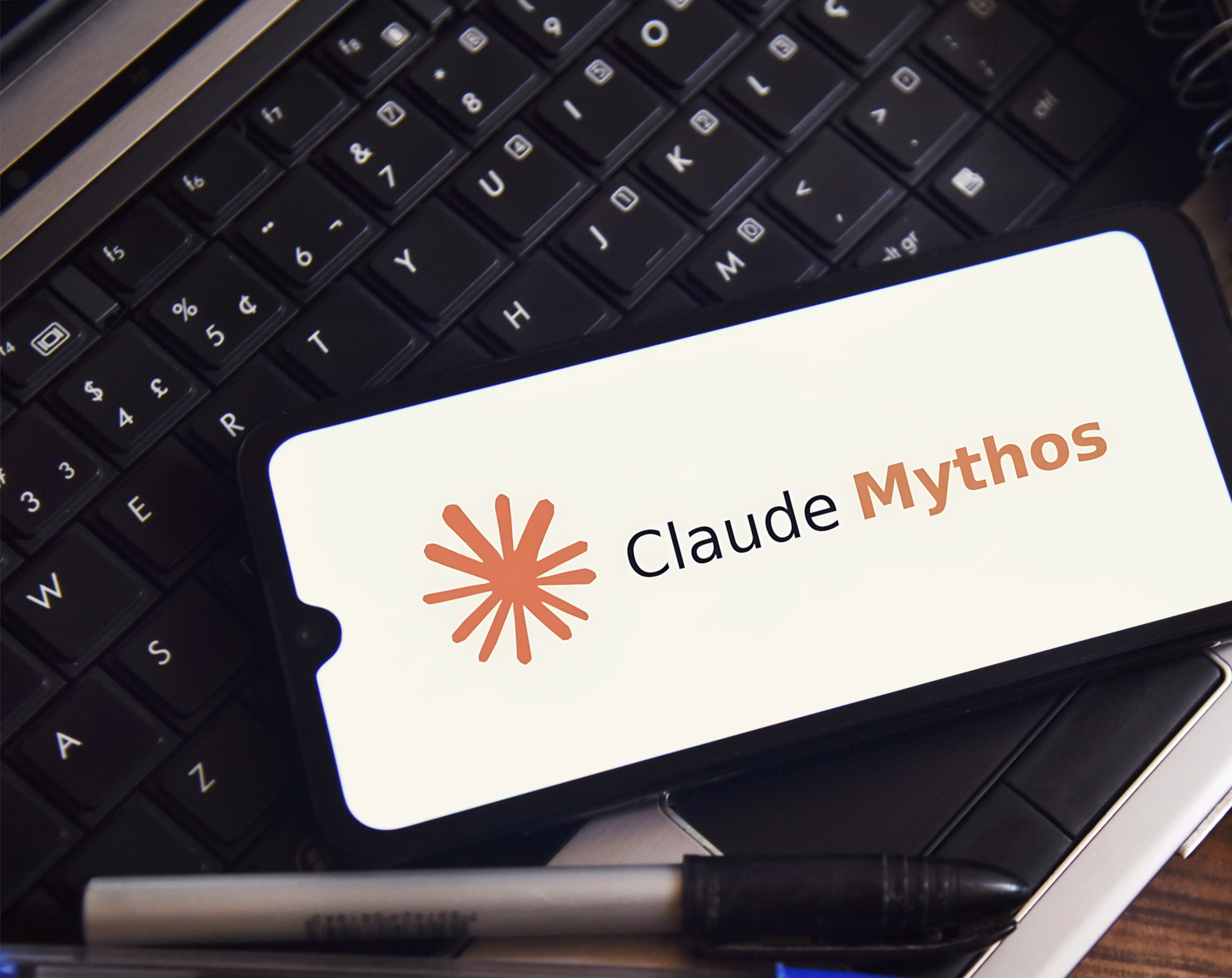 The Claude Mythos logo is displayed on a smartphone screen on a table with office objects. Concept of technology, computing, and innovation.